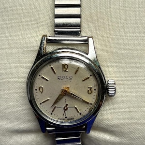 Può includere: Un orologio da polso ROCO vintage con cinturino e cassa in metallo argentato. Il quadrante dell'orologio è color crema con lancette e indici dorati. Il testo "ROCO" è stampato sopra il centro e "SWISS MADE" è stampato in basso.