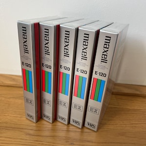 Peut inclure: Une pile de cinq cassettes vidéo VHS Maxell E-120. Les cassettes ont un emballage argenté avec des rayures verticales rouges, vertes et bleues. La marque "maxell" est imprimée en noir en haut.