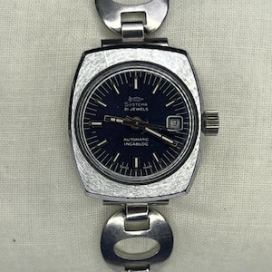 Könnte beinhalten: Eine Vintage-Systema-Armbanduhr mit dunkelblauem Zifferblatt und silberfarbenem Metallarmband. Das Zifferblatt zeigt den Text "Systema 21 Jewels" und "Automatic Incabloc". Die Uhr hat eine Datumsanzeige bei 3 Uhr.