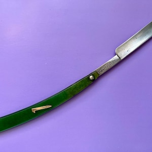 Op de afbeelding: Een vintage recht scheermes met een zilveren lemmet en een gebogen groene handgreep. De handgreep heeft een goudkleurig metalen accent. Het lemmet heeft geëtste tekst. Het scheermes ligt op een paarse achtergrond.
