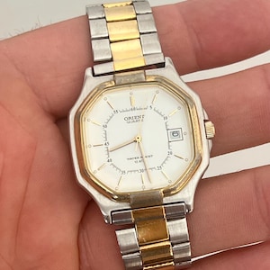 Op de afbeelding: Een Orient Quartz polshorloge met een witte achthoekige wijzerplaat, goudkleurige ring en een tweekleurige schakelarmband van zilver- en goudkleurig roestvrij staal. De wijzerplaat heeft goudkleurige wijzers, een datumvenster en markeringen.