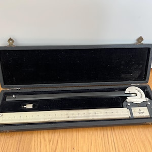 Puede incluir: Un planímetro vintage A. Ott Wien alojado en un estuche rectangular negro con interior de terciopelo negro. El instrumento de precisión cuenta con una regla metálica con marcas hasta 23 cm, un brazo negro y un dial circular con piezas.