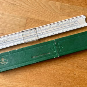 Può includere: Un regolo calcolatore bianco Faber-Castell con un cursore in plastica trasparente e una custodia protettiva verde testurizzata. La custodia presenta il logo Faber-Castell e la scritta "CASTELL-ELEKTRO 1/92" in rilievo sulla parte anteriore.