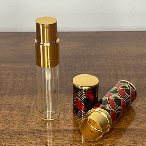 Puede incluir: Un frasco de perfume de vidrio transparente con detalles dorados y el texto "Nuelle". El frasco está junto a una tapa dorada y un estuche decorativo con un diseño art déco negro, rojo y dorado.