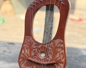 May include: A handcrafted, wooden lyre with a rich, reddish-brown finish. The instrument features intricate floral carvings on the body and a series of strings stretched across the sound hole. The lyre is leaning against a wooden post.