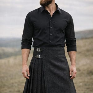 Puede incluir: Un hombre con una camisa negra y un kilt gris. El kilt tiene pliegues y hebillas. La tela parece ser lana texturizada. El fondo es una escena borrosa al aire libre.