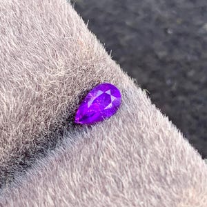 May include: A faceted, pear-shaped, vibrant purple gemstone. The gem is set against a soft, textured, gray background. The cut of the stone creates multiple facets that reflect light, enhancing its brilliance. This gemstone is suitable for jewelry making.