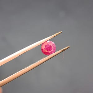 May include: A round, faceted ruby gemstone held by tweezers. The ruby is a vibrant red color, and the tweezers are a light gold color. The background is a solid gray.