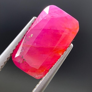 Könnte beinhalten: Ein facettierter Edelstein in rechteckiger Form, gehalten von einer silbernen Pinzette. Der Edelstein zeigt einen Farbverlauf von Rosa und Rot, mit polierter Oberfläche und sichtbarer innerer Struktur. Der Hintergrund ist dunkelgrau.