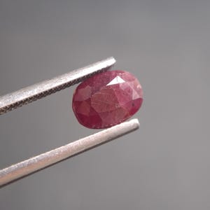 May include: A faceted, oval-shaped ruby gemstone held by silver tweezers. The ruby is a deep red colour with multiple facets that reflect light. The gemstone is a natural, untreated ruby.