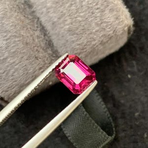 Puede incluir: Una gema rosa, de corte rectangular, es sostenida por unas pinzas plateadas sobre un fondo gris y negro. La gema tiene un interior brillante y reflectante y un tono rosa intenso, mostrando sus facetas.