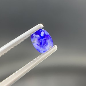 May include: A small, oval-shaped, translucent blue gemstone with visible internal inclusions and a polished surface. The stone displays varying shades of deep and light blue, featuring a mottled texture throughout its structure, held by metal tweezers.
