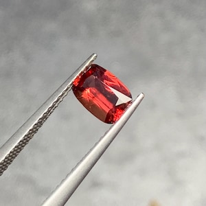 Könnte beinhalten: Ein kissenförmig geschliffener Edelstein mit tiefroter Farbe und facettierter Oberfläche, gehalten von einer silberfarbenen Metallpinzette. Der Stein zeigt innere Reinheit und Lichtreflexion auf seinen polierten Facetten und präsentiert einen lebendigen, gesättigten Karmesinton.