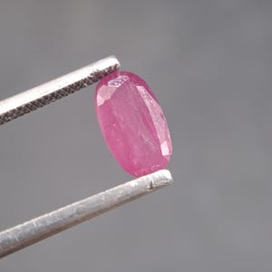 May include: A faceted, oval-shaped ruby gemstone held by silver tweezers. The ruby is a vibrant pinkish-red color, with visible facets catching the light. The background is a solid, neutral gray.