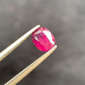 May include: A faceted, oval-shaped ruby gemstone held by tweezers. The ruby is a vibrant, deep red color with visible internal inclusions. The tweezers are silver-colored, and the background is a blurred dark gray.