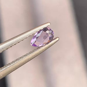 May include: A pear-shaped, faceted amethyst gemstone held by tweezers. The gemstone is a vibrant purple color and has a polished surface. The tweezers are silver-colored and have a textured grip.