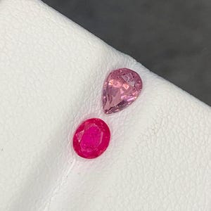 May include: Two loose gemstones: one pear-shaped light pink stone with a faceted surface and one oval-shaped deep red stone with a faceted surface. Both stones are displayed on a white textured surface with a visible crease running between them.