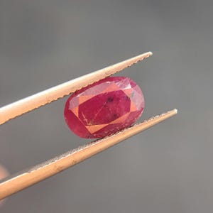 May include: A faceted, oval-shaped ruby gemstone held by tweezers. The ruby is a deep red color with internal inclusions and a polished surface. The tweezers are a copper color, and the background is a solid gray.