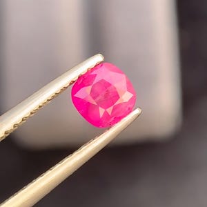 Peut inclure: Une pierre précieuse rose vif à facettes est tenue par une pince. La gemme est de taille coussin, avec de multiples facettes réfléchissant la lumière. La pince est de couleur or clair, et l'arrière-plan est un flou gris et noir.