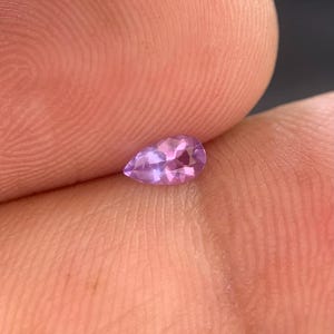 May include: A small, pear-shaped gemstone with a faceted surface and a vibrant, translucent purple hue. The stone displays internal light reflections and a clear, polished appearance, showcasing a distinct teardrop silhouette with a pointed top.