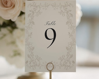 May include: A table number card with the number 9 in black, displayed on a gold-colored stand. The card has a floral border and the word "Table" above the number. The background includes flowers and a candle.
