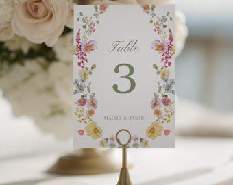 May include: A wedding table number card featuring the number 3 in green, the word "Table" above, and names "Maisie & Jamie" below. The card is held in a gold metal stand and decorated with colorful flowers.
