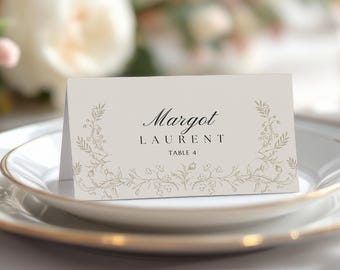 May include: A formal place setting with a white plate and gold rim, holding a folded place card. The card is beige with the name "Margot Laurent" and "Table 4" printed on it, with floral detailing.