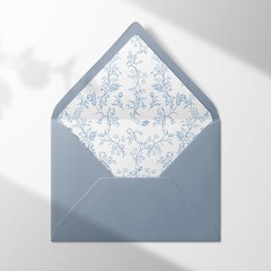 Könnte beinhalten: Ein staubblauer Umschlag mit spitzer Klappe und einem Innenfutter, das mit einem zarten blau-weißen botanischen Blumenmuster verziert ist. Der Umschlag ist geöffnet und gibt den Blick auf das komplexe, sich wiederholende Ranken- und Blattmuster frei.