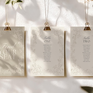 Op de afbeelding: Drie ivoorkleurige trouwzitkaarten, opgehangen met gouden clips. De linkerkaart leest "Find Your Seat". De middelste en rechterkaarten zijn gelabeld met "Table One" en "Table Two", met gastennamen. Bloemenranden omlijsten de tekst.