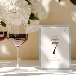 May include: A table number card with a light blue border displays "Table 7" in elegant script, with the names "Rosie & Marcus" below. Two wine glasses filled with red wine are in the background, with white flowers.