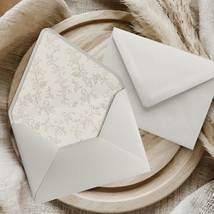 Peut inclure: Deux enveloppes blanches, dont une ouverte révélant un intérieur à motifs floraux, reposent sur une assiette en bois. L'enveloppe ouverte présente un délicat motif floral beige. La seconde enveloppe est fermée. Le fond présente un tissu aux tons neutres et des plantes séchées.