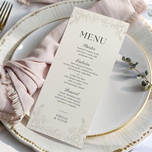 Może przedstawiać: Karta menu weselnego z kwiatowym wzorem, z napisem "MENU" na górze. Menu zawiera przystawkę, dania główne i deser. Nazwiska "AMELIA & NOAH" znajdują się na dole. Karta znajduje się na białym talerzu ze złotą obwódką, z różową serwetką.