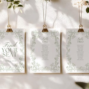 Puede incluir: Tres tarjetas de plano de mesa de boda suspendidas por clips dorados. Cada tarjeta presenta un borde floral y texto: "Find Your Seat", "Table One" y "Table Two". Las tarjetas son blancas con texto verde.