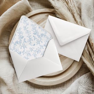 May include: Two white envelopes, one open, revealing a blue floral patterned interior. The envelopes rest on a wooden plate, with dried grasses and a beige fabric in the background. The open envelope displays a delicate blue floral design.