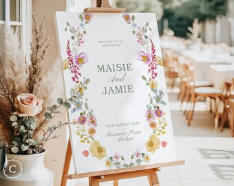 Pode incluir: Uma placa de boas-vindas de casamento em um cavalete de madeira. Apresenta os nomes "Maisie e Jamie" e a data "10 de agosto de 2028". Uma grinalda floral em rosa, amarelo e azul envolve o texto. Um vaso com flores está em primeiro plano.