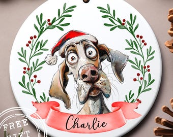 May include: A white ceramic Christmas ornament featuring a cartoon dog wearing a Santa hat. The dog's name, "Charlie," is on a red banner. Green leaves and red berries form a wreath around the dog.