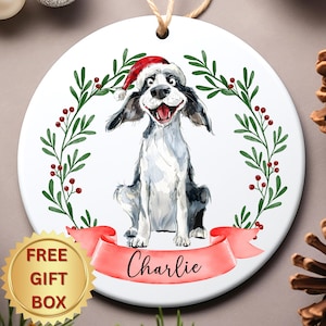 May include: A round, white Christmas ornament featuring a watercolor illustration of a dog wearing a Santa hat. The dog is surrounded by a green wreath with red berries and a red banner with the name "Charlie". A gold "FREE GIFT BOX" sticker is in the lower left.