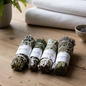 Puede incluir: Cuatro manojos de hierbas secas, incluyendo salvia, enebro, eucalipto y lavanda, se exhiben sobre una superficie de madera. Cada manojo está envuelto con hilo y etiquetado. Toallas blancas y un cuenco pequeño en el fondo.