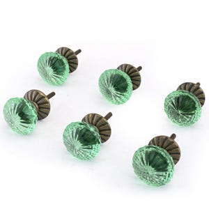 May include: A set of six round cabinet knobs featuring translucent light green glass with a faceted, sunburst-like pattern. Each knob is attached to a circular, dark bronze-toned metal base with a threaded screw extending from the back.
