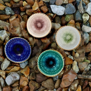May include: Four small, decorative ceramic bowls in shades of blue, teal, pink, and green, each with a gold rim and intricate floral patterns. The bowls are arranged on a bed of pebbles and stones, creating a natural and artistic display.