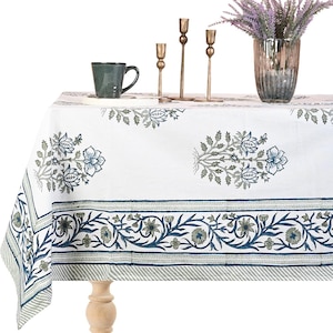 May include: White tablecloth with a blue and green floral pattern. The tablecloth has a border with a repeating floral design. A green mug, brass candlesticks, and a vase with purple flowers are on the table.