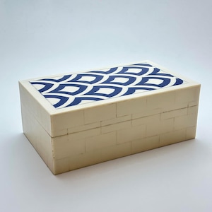 Puede incluir: Caja rectangular de color crema con un patrón geométrico azul y blanco en la tapa. Los lados de la caja tienen un diseño similar a ladrillos. Un artículo de almacenamiento decorativo.