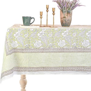 Puede incluir: Un mantel verde claro con un estampado floral y paisley blanco. El mantel tiene un borde marrón y un diseño de chevrón. Una taza verde, candelabros dorados y un jarrón con flores moradas están sobre la mesa.