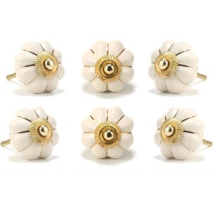 Może przedstawiać: Sześć białych gałek do szafek w kształcie kwiatów ze złotymi akcentami. Gałki mają falisty brzeg i centralne złote kółko. Okucia przeznaczone są do mebli i dekoracji wnętrz.