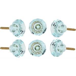 Puede incluir: Seis pomos de armario de cristal transparente facetado con un tinte azul claro. Cada pomo tiene un tornillo dorado y un centro decorativo. Los pomos están dispuestos sobre un fondo blanco, mostrando sus cualidades reflectantes.