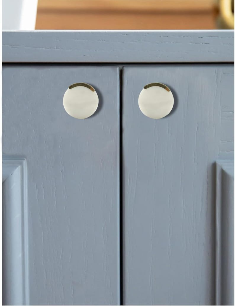 May include: Close-up of a light blue cabinet with two white and gold cabinet knobs. The knobs are round with a gold trim and a white center. The cabinet doors have a recessed panel design.