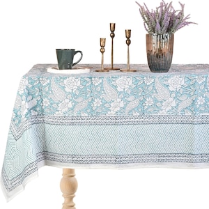 May include: A rectangular tablecloth with a light blue and white floral paisley design. The tablecloth is draped over a wooden table, with a green mug, brass candle holders, and a vase of purple flowers on top.