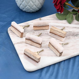 May include: Six rectangular cabinet knobs with a decorative design. Each knob combines light wood, a white stone-like material, and a brass-coloured screw. The knobs are arranged on a white marble surface.