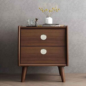 May include: A mid-century modern style nightstand with two drawers. The nightstand is made of dark brown wood and features two round, silver drawer pulls. The legs are angled and tapered. A small vase with yellow flowers sits on top.