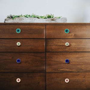 May include: A dark brown wooden dresser with six drawers, each adorned with a unique, colorful knob. The knobs feature shades of teal, green, blue, and pink, adding a pop of color to the furniture. A rectangular planter with greenery sits on top.